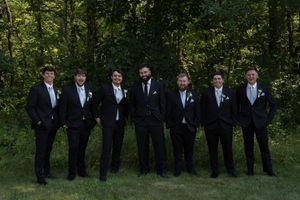 A line of groomsmen in black tuxedos pose together outdoors against a natural backdrop.