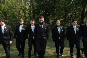 Wedding party members in formal black attire pose together on grass with trees in background.