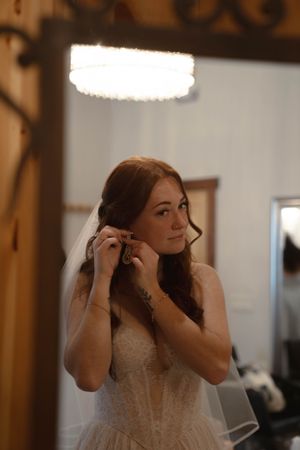 A bride in a white lace dress adjusts her veil in an indoor setting.
