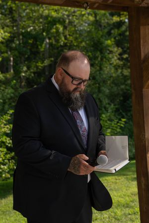 A man in a black suit reads from a book under a wooden pergola.