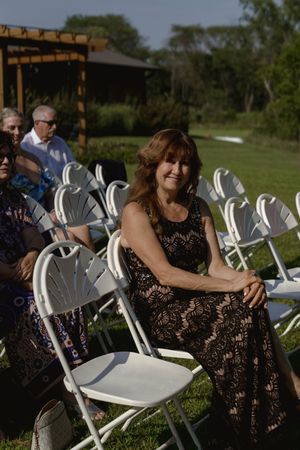 A guest in a black lace dress sits among white folding chairs at an outdoor wedding.