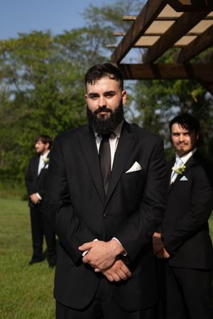 Groomsmen in black suits and ties stand in formation during an outdoor ceremony.