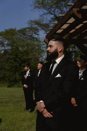 Groomsmen in black tuxedos await the start of an outdoor wedding ceremony.