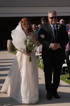 A bride in a white lace dress walks down a pathway at an outdoor wedding ceremony.