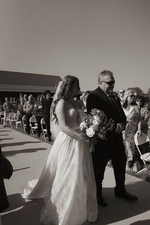 Bride in flowing white gown walks with guests at outdoor wedding reception.