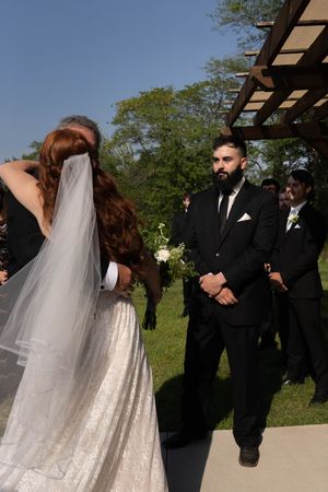 A couple stands together during their outdoor wedding ceremony under a wooden pergola.