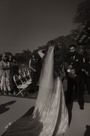 Wedding party and guests gather outdoors with white chairs visible in background.