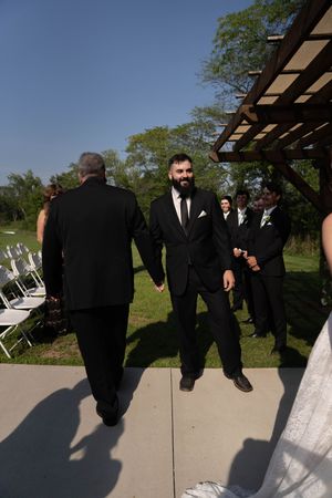 Wedding party members in black suits stand on a concrete path during the ceremony.