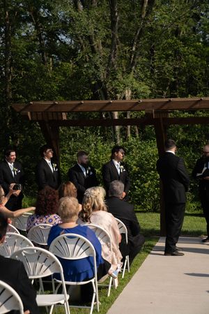 Wedding guests seated in white chairs beneath a wooden pergola during an outdoor ceremony.