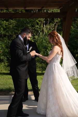 Wedding ceremony moment as couple exchanges rings beneath a rustic pergola in a garden setting.