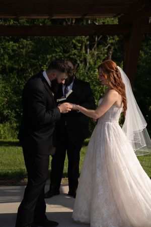 A couple exchanges rings during an outdoor wedding ceremony under a wooden pergola.