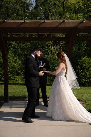 Wedding ceremony taking place under a wooden pergola with lush greenery in the background.