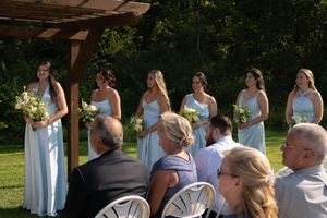 Bridesmaids in light blue dresses stand at an outdoor wedding ceremony with guests seated.