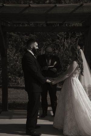 Wedding ceremony taking place under wooden pergola with couple exchanging vows.
