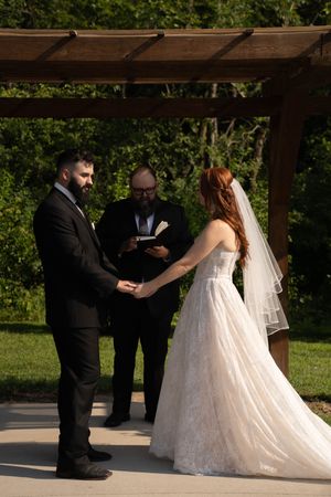 Outdoor wedding ceremony with couple and officiant standing under a wooden pergola structure.