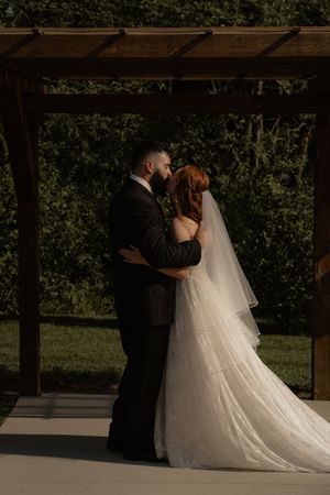 A couple embraces during their wedding ceremony under a rustic wooden pergola.