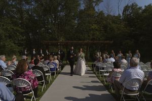 Outdoor wedding ceremony setup with white chairs arranged in rows under trees and a wooden pergola.