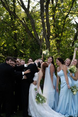 Wedding party in light blue bridesmaid dresses and black suits poses playfully outdoors under green trees.