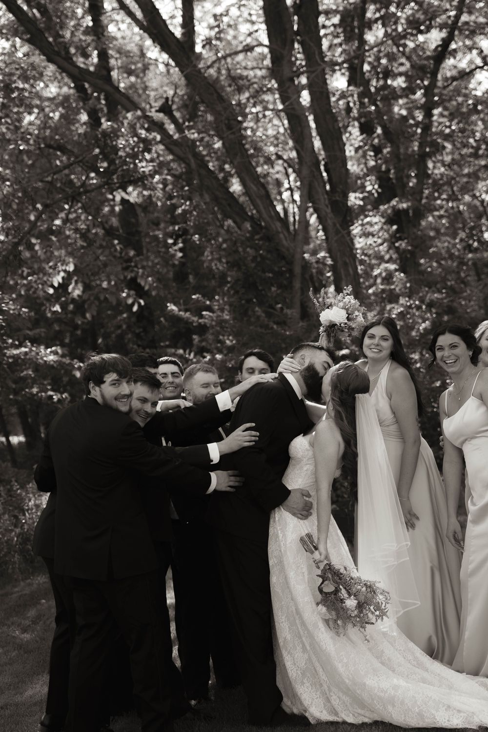Wedding party poses together outdoors with bridesmaids in light dresses and groomsmen in dark suits.