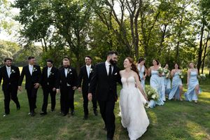 Wedding party in formal attire walks across a lush green lawn with trees in the background.