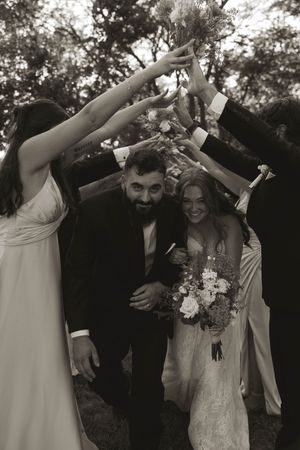 Wedding party celebrates with raised arms and bouquets in black and white photo.