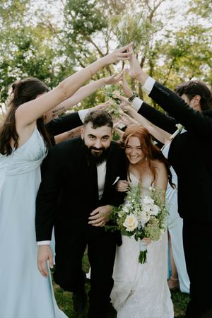 Wedding party forms a tunnel with raised arms while couple walks underneath with bouquet.