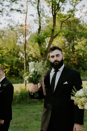 Groomsman in black tuxedo holds delicate white and green floral bouquet outdoors.