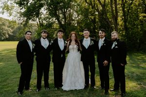Wedding party in formal black tuxedos and white gown pose together on green lawn.