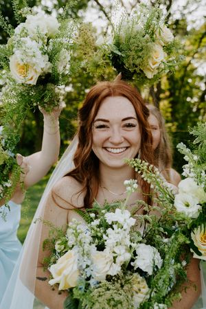 Elegant bridal portrait surrounded by lush white floral arrangements and greenery in natural outdoor setting.