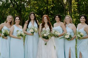 Bridesmaids in pale blue dresses pose with bouquets against green outdoor backdrop.