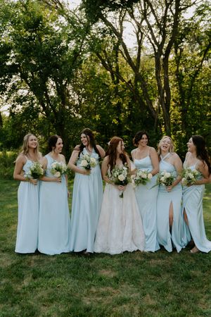 Wedding party in light blue bridesmaid dresses posing together on grassy hillside with trees.