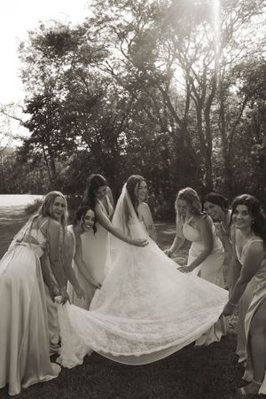Black and white photo of bridal party arranging wedding dress train in outdoor setting.