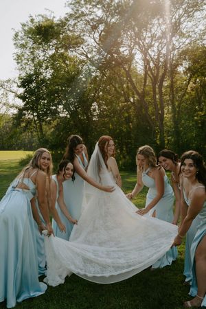Bridesmaids in light blue dresses helping arrange wedding dress train at sunset.