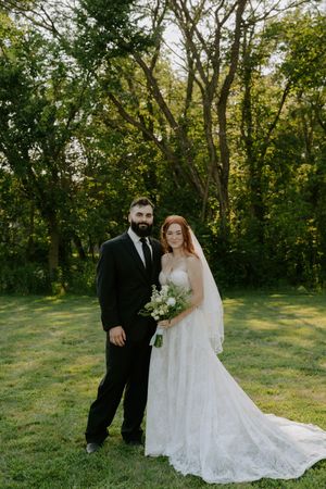 A wedding couple poses together on a grassy hill with trees in the background during golden hour.