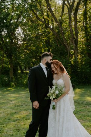 A romantic wedding portrait of a couple in formal attire standing among trees at sunset.
