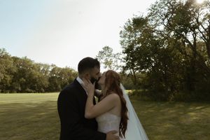A couple shares their first kiss as newlyweds in a sunlit outdoor field surrounded by trees.