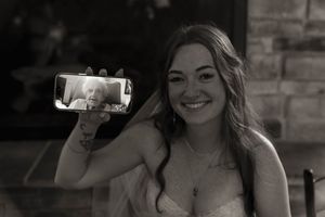 A bride in a white dress holds up a phone showing a video call during the wedding celebration.