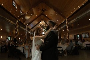 A romantic first dance takes place under elegant chandeliers and string lights in a rustic wooden reception venue.