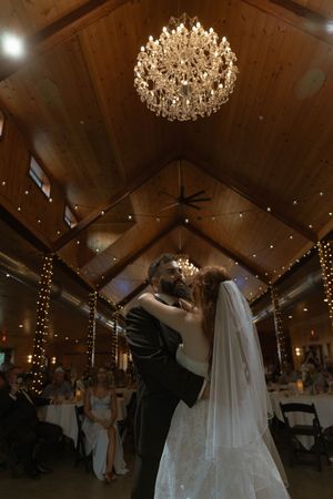 A couple shares an intimate dance moment beneath sparkling chandeliers in a warmly lit barn venue.