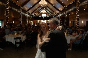 Wedding guests celebrate on the dance floor of a rustic venue decorated with string lights and chandeliers.