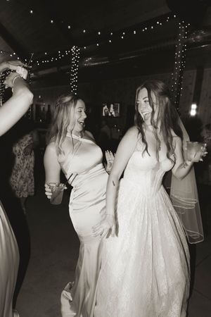 Wedding guests in light-colored dresses dance beneath twinkling fairy lights at an evening reception.