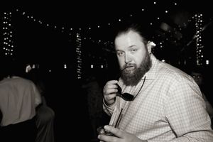 Black and white portrait of a bearded guest adjusting his bowtie at a wedding reception.