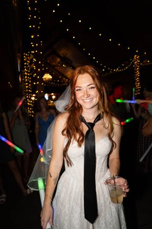 Bride wearing a white dress and black tie smiling at a reception with string lights.