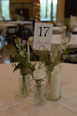 Wedding table centerpiece with table number 17, white flowers in crystal vases, and a white candle on a white tablecloth.