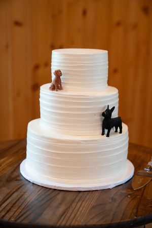 Three-tiered white wedding cake decorated with silhouettes of two dogs on wooden table.