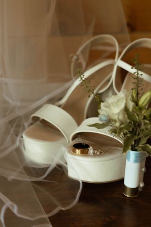 White platform wedding shoes with veil and floral arrangement on dark wooden surface.