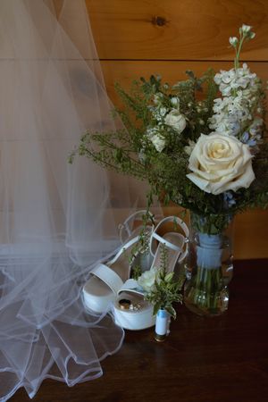 White bridal shoes with veil and floral arrangement featuring roses against wooden backdrop.