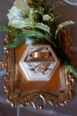 Wedding rings displayed in hexagonal box with floral arrangement on ornate gold tray.