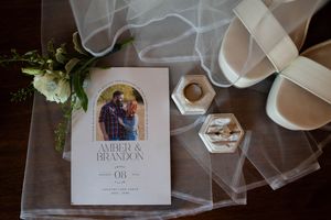 Wedding invitation, rings in hexagonal box, and white shoes arranged with veil on wooden surface.
