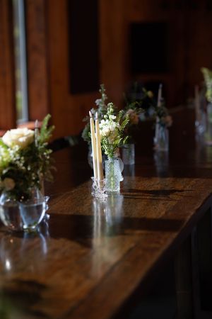 Multiple glass vases with white and green floral arrangements line a rustic wooden table.